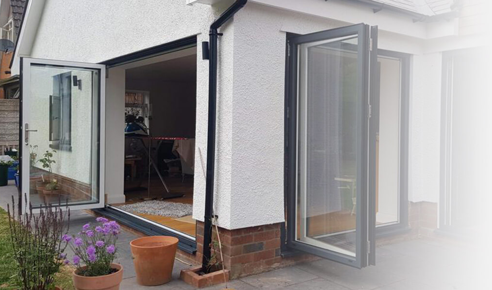 A partially open aluminium bi-fold door in a traditional white home, leading to a cozy and inviting interior living space, with potted plants adding a touch of greenery outside.