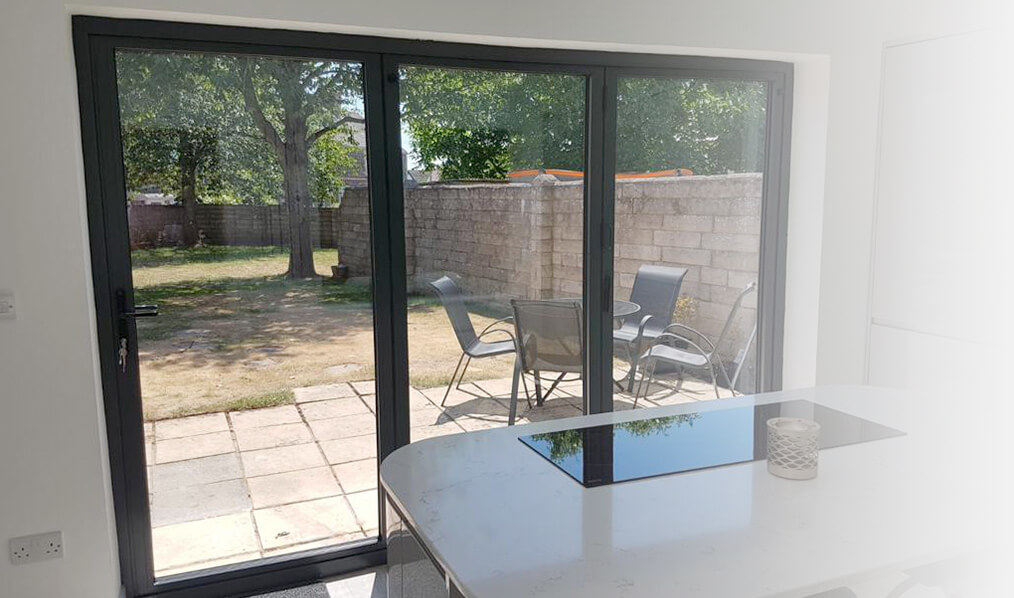Interior view of a modern home featuring a black aluminium bi-fold door leading to a patio with a garden view, complemented by a sleek white dining table.