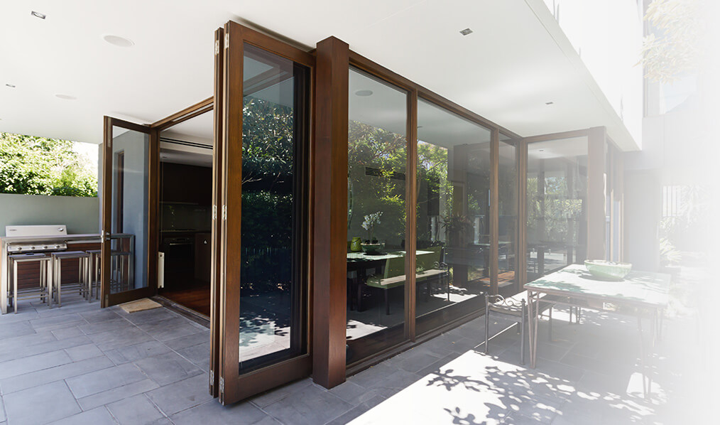 Open brown aluminium bi-fold doors connecting a luxurious indoor kitchen and dining space with an outdoor patio, enhancing the flow of natural light and indoor-outdoor living.
